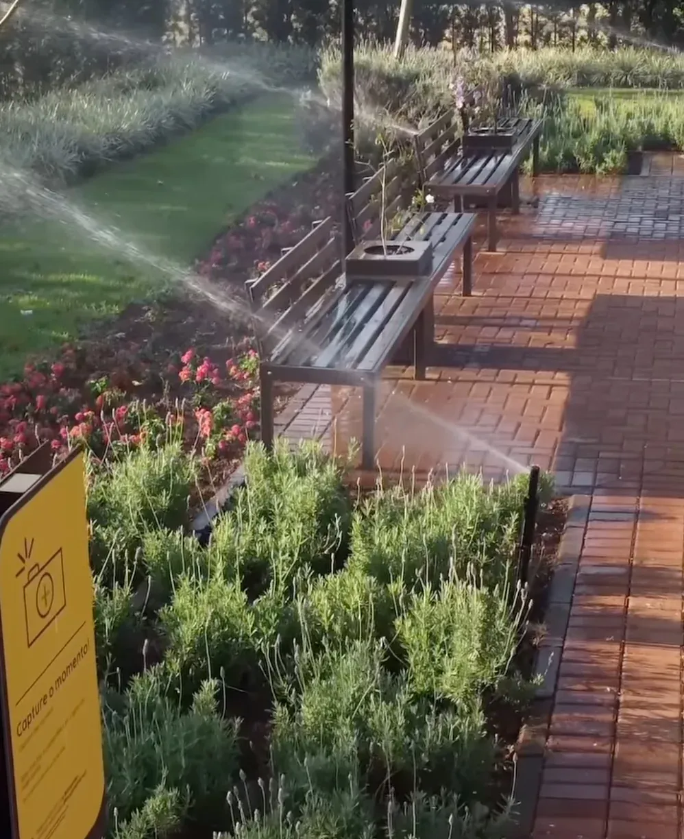 Área de convivência em jardim com aspersores irrigando canteiros